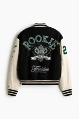 Campera estilo varsity con cuerpo negro y mangas en contraste color crema. Presenta múltiples parches y bordados decorativos en el frente y las mangas, cuello, puños y ruedo acanalados a rayas, y cierre frontal con botones a presión.