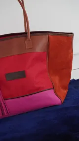 Bolso de fin de semana tipo tote, confeccionado en una combinación de lona y gamuza en tonos rojo, fucsia y marrón. Cuenta con doble asa de hombro, cierre superior y detalle de borlas colgantes en el lateral.