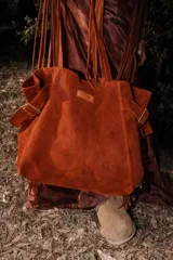 Bolso tipo shopper de cuero de descarne color terracota, con asas de mano y detalles de hebillas metálicas doradas en los laterales. Presenta flecos colgantes en la parte inferior y un parche de cuero grabado en el frente.