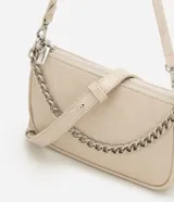 Cartera de mano tipo baguette, de color beige, con cierre superior de cremallera y una cadena decorativa plateada en la parte frontal. Cuenta con una correa larga ajustable y extraíble para usar al hombro o cruzada.
