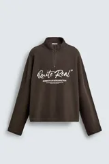 Sudadera de corte boxy fit en tejido de algodón con textura acanalada, color marrón oscuro. Presenta cuello subido con cierre frontal de cremallera y estampado frontal con texto en contraste.