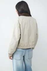 Campera acolchada corta color beige, con cuello de pana en contraste color marrón. Cuenta con cierre frontal de cremallera y botones a presión, bolsillos laterales, y puños y bajo elásticos.