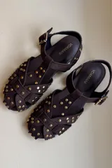 Sandalia tipo guillermina confeccionada en cuero nobuk color chocolate, con tiras entrelazadas en la parte delantera y tachas doradas. Cuenta con una tira en el tobillo con hebilla dorada.