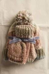 Set de invierno compuesto por un gorro con pompón y un par de guantes, ambos de tejido de punto grueso con efecto jaspeado en tonos tierra (marrón, beige, verde oliva). El set está atado con una cinta de terciopelo gris.