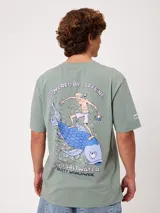 Remera de manga corta color verde militar, corte holgado (loose fit), con estampa artística en la espalda que muestra a un personaje surfeando sobre un pez grande y texto descriptivo.
