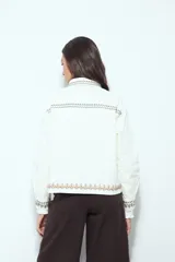 Campera de jean color blanco con bordados geométricos en cuello, delantero, bolsillos, puños y ruedo. Tiene botones dorados y un calce recto y relajado.