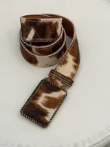 Cinto de cuero vacuno con pelo, estampado animal print tipo vaca en tonos marrones y blancos, con hebilla rectangular forrada en el mismo material.