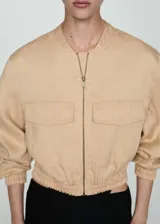 Chaqueta tipo bomber color beige, con cierre frontal de cremallera, cuello redondo, bolsillos de solapa en el pecho y cintura y puños elásticos.