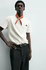 Camiseta de corte relajado (relaxed fit) color blanco hueso, con cuello redondo, manga corta y bolsillo plastrón en el pecho. Presenta un bordado pequeño y contrastante de una palmera en el área del bolsillo.