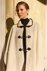 Capa color crudo con cuello camisero y ribetes negros. Cierre frontal con alamares negros y bolsillos laterales.