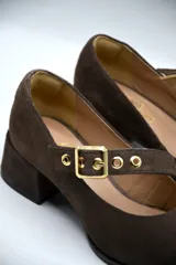 Zapato tipo Mary Jane de gamuza color marrón, con taco cuadrado medio y tira ajustable en el empeine con hebilla metálica dorada y detalles de ojalillos.