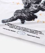 Remera blanca de algodón, corte oversized, con cuello redondo y mangas cortas. Presenta un estampado frontal con un ángel y el texto "PARADISE" en negro, y un pequeño gráfico de estrella/brújula en el lado izquierdo del pecho.