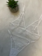 Conjunto de lencería de encaje blanco compuesto por un bralette triangular, una tanga con tiras laterales y un portaligas integrado. El diseño presenta detalles de encaje floral y breteles regulables.