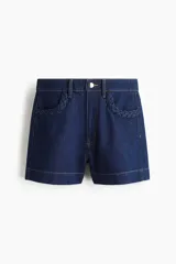 Shorts de denim azul oscuro, tiro alto, con cierre de botón dorado. Los bolsillos delanteros tienen un detalle de ribete trenzado.