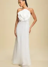 Vestido largo de fiesta color blanco, con escote halter y un gran aplique floral tridimensional en el pecho. Presenta una falda fluida con caída suave y cierre posterior.