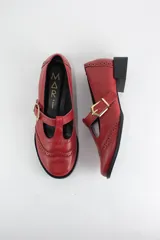 Zapato tipo guillermina estilo oxford, confeccionado en cuero negro con detalles de perforaciones decorativas en la puntera y cierre mediante tira con hebilla metálica dorada. Presenta un taco bajo cuadrado.