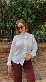 Camisa blanca de textura sutil, con cuello alto tipo victoriano y detalle de volados asimétricos a lo largo de la botonadura frontal. Presenta mangas largas con puños terminados en volados.