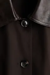Abrigo oversized de sarga color marrón con cuello de cuero sintético. Modelo con botones en el frente, bolsillos laterales discretos, mangas tipo capa y aberturas laterales con botón.