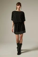 Vestido corto de poliéster color negro con escote a la base, mangas cortas y diseño holgado. Cuenta con detalles de tachas metálicas plateadas distribuidas por la prenda y agrupadas en el dobladillo y los puños.
