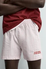 Bóxer tipo short de popelín con estampado de rayas verticales finas en blanco y rosado/rojo. Posee cintura elástica y una pequeña inscripción bordada en rojo en la parte inferior de una de las piernas.