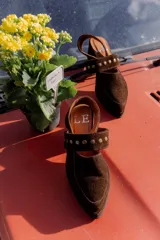 Zuecos de gamuza color marrón con taco cuadrado bajo y tira sobre el empeine decorada con tachas metálicas.