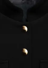 Chaqueta de estilo militar en color negro, con cuello redondo, manga larga y cierre frontal de botones metálicos dorados. Presenta cuatro bolsillos de solapa en la parte delantera y diseño de corte recto.