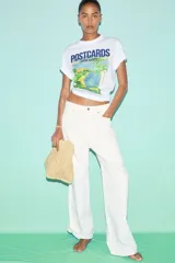 Remera blanca de manga corta y cuello redondo, con un estampado gráfico que simula una postal de Brasil, mostrando una playa tropical con palmeras y vegetación.