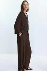 Pantalón negro de tiro alto con cintura elástica ajustable mediante cordones con terminales dorados. Presenta pernera ancha y bolsillos laterales. La tela tiene una textura sutilmente arrugada o plisada.