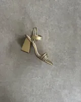 Sandalia de fiesta dorada con tiras finas cruzadas en el empeine, pulsera con hebilla al tobillo y taco geométrico.