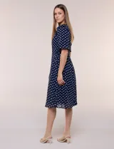 Vestido midi azul marino con estampado de lunares blancos, confeccionado en gasa semitraslúcida. Tiene escote redondo, mangas cortas acampanadas con volantes en los hombros, lazo ajustable en la cintura y falda con vuelo y forro interior.
