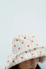 Gorro tipo bucket de algodón, color crudo, con rayas verticales finas en tono neutro y pequeños corazones rojos bordados distribuidos por toda la superficie. Presenta ala ancha con terminación deshilachada.