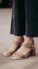 Sandalia de cuero color taupe con taco cuadrado de 6 cm, tira frontal ancha y tira en el tobillo con detalle metálico dorado y hebilla circular.