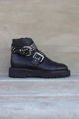 Borcego negro de cuero con plataforma y suela track. Presenta dos correas con hebillas plateadas y apliques de tachas redondas.