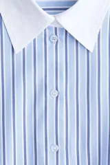 Camisa oversized de popelina de algodón con diseño a rayas verticales en tonos azul y blanco. Presenta cuello clásico y puños anchos en color blanco sólido, cierre frontal con botones, hombros caídos y bajo redondeado con aberturas laterales.