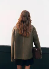 Abrigo de corte recto y estilo clásico en color verde oliva, confeccionado con efecto piel y forro interior de borreguito. Presenta cuello camisero, cierre frontal con botones y dos bolsillos laterales.