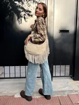 Cartera tipo hobo de cuero color arena, con diseño de media luna y flecos largos en la parte inferior. Cuenta con correa ajustable para llevar al hombro y cierre superior.