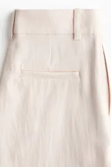 Pantalón sastrero de tiro alto en mezcla de lino y viscosa, de corte amplio y perneras rectas con pliegues frontales. Cuenta con trabillas para cinturón, cierre frontal con cremallera y corchete, y bolsillos laterales discretos.