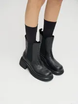 Bota tipo Chelsea de caña media, confeccionada en cuero vacuno negro con acabado mate. Presenta paneles laterales elásticos, tirador trasero y una suela robusta con plataforma delantera y taco cuadrado.