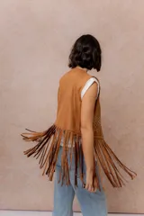 Chaleco de gamuza sintética color tostado, con diseño abierto sin cierre. Presenta bordados decorativos en hilo blanco y flecos largos en el delantero y la espalda.