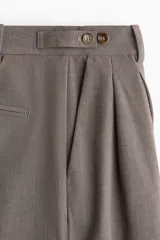 Pantalón sastrero de corte recto y perneras amplias, color marrón, con pinzas frontales, trabillas ajustables con botones en la cintura y raya marcada en el centro de las piernas.