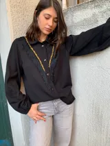 Camisa negra de tejido fino con cuello clásico, cierre frontal con botones, mangas largas abullonadas con puños elásticos y detalle de galón dorado y flecos negros en el frente.