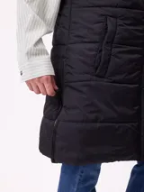 Chaleco largo acolchado color beige, con capucha forrada en sherpa, cierre frontal con cremallera y botones a presión, y bolsillos laterales.