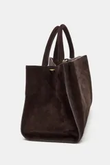 Bolso de mano formato city confeccionado en gamuza color marrón oscuro. Cuenta con doble asa de mano, asa bandolera extraíble, compartimento interior con cierre y bolsillo exterior con imán.