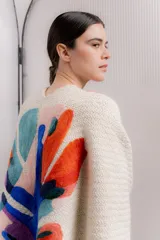 Ruana de tejido de punto grueso en color crudo, con un diseño artístico bordado en la espalda que presenta formas orgánicas y abstractas en colores vibrantes como naranja, azul, turquesa y violeta.