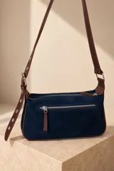 Cartera tipo bandolera de gamuza marrón, con detalles de tachas y ojalillos metálicos en el frente. Cuenta con cierre superior, bolsillo trasero con cierre, correa ajustable y placa metálica con la marca grabada.
