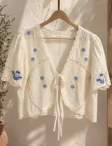 Camisa blanca de manga corta con bordados florales en azul, escote en V con detalle de lazos para atar en el frente y terminaciones de encaje.