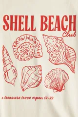 Remera de algodón de corte holgado en color crema, con cuello redondo y mangas cortas. Presenta un estampado frontal en color rojo con ilustraciones de caracoles de mar y el texto 'SHELL BEACH Club'.