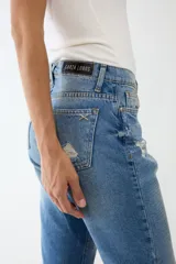 Jean de algodón 100% con lavado azul medio, tiro alto y corte recto/acampanado. Presenta un estampado texturizado tipo cocodrilo en tono más claro y detalles de desgaste/roturas.