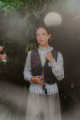 Chaleco de algodón color marrón oscuro con bordados en contraste de colores vibrantes como azul, amarillo, verde, rosa y blanco. Tiene bordes festoneados en azul y amarillo, y tiras para atar en el frente.