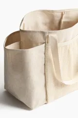 Cartera shopper grande confeccionada en tejido grueso de mezcla de yute color beige, con doble asa de hombro, compartimentos de malla en el frente y dorso, y bolsillos laterales abiertos.